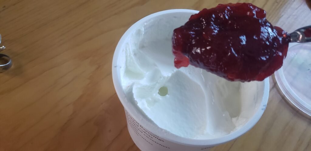 Spoonful of cold skyr and Bonne Maman strawberry preserves over an open tub.