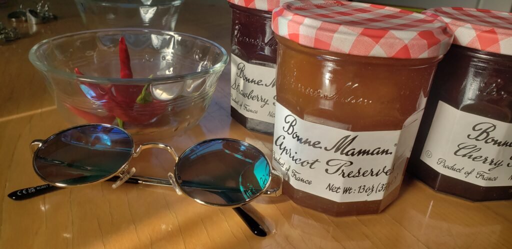 Three jars of Bonne Maman preserves on a wood table — strawberry, apricot, and cherry — with round sunglasses.